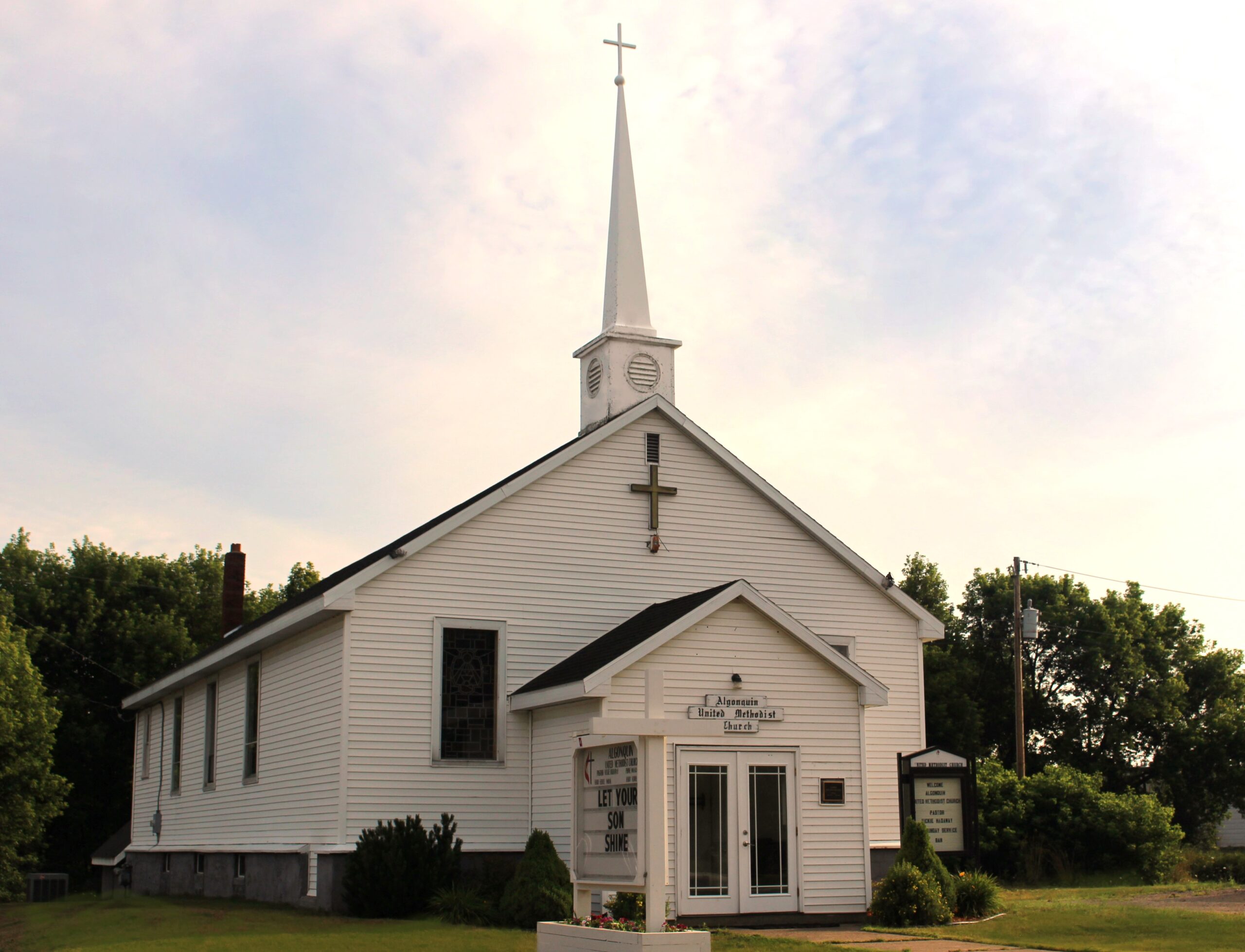 White clapboard church with a tall steeple and cross, featuring a sign that invites community engagement and services.