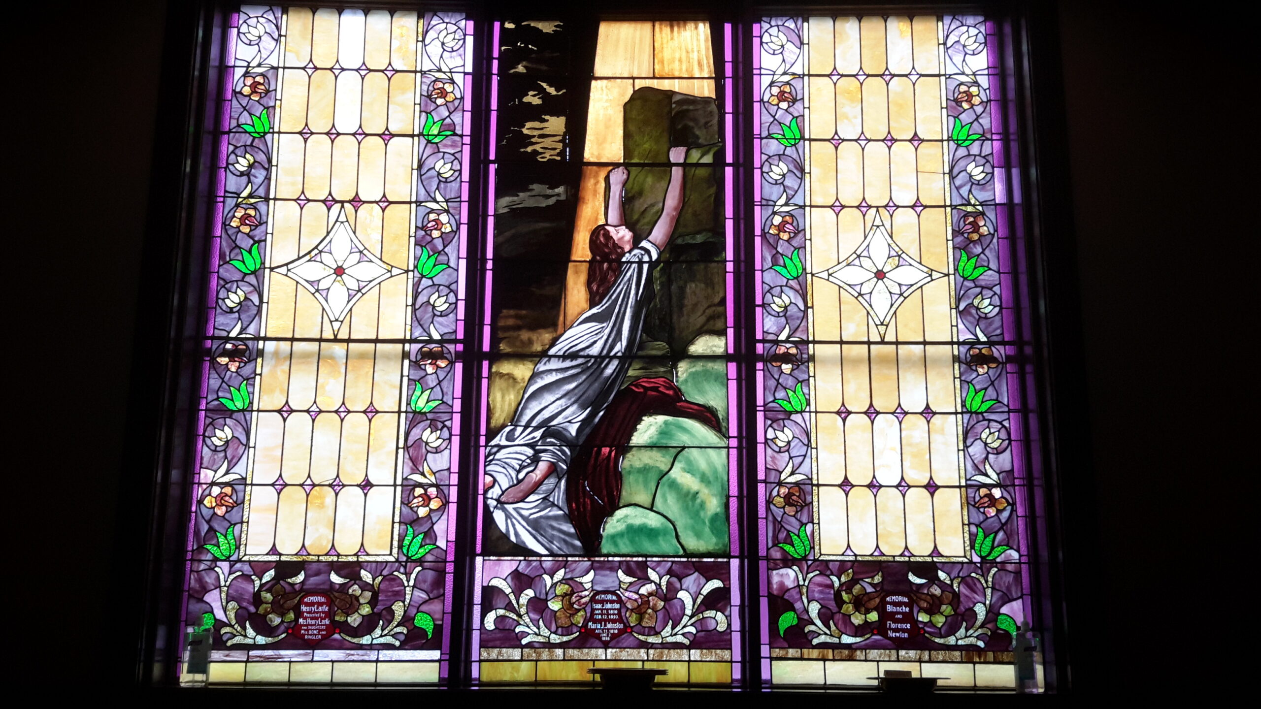 Stained glass window depicting a figure reaching upwards, surrounded by ornate floral patterns and colorful glass. Highlights artistic craftsmanship in religious settings.